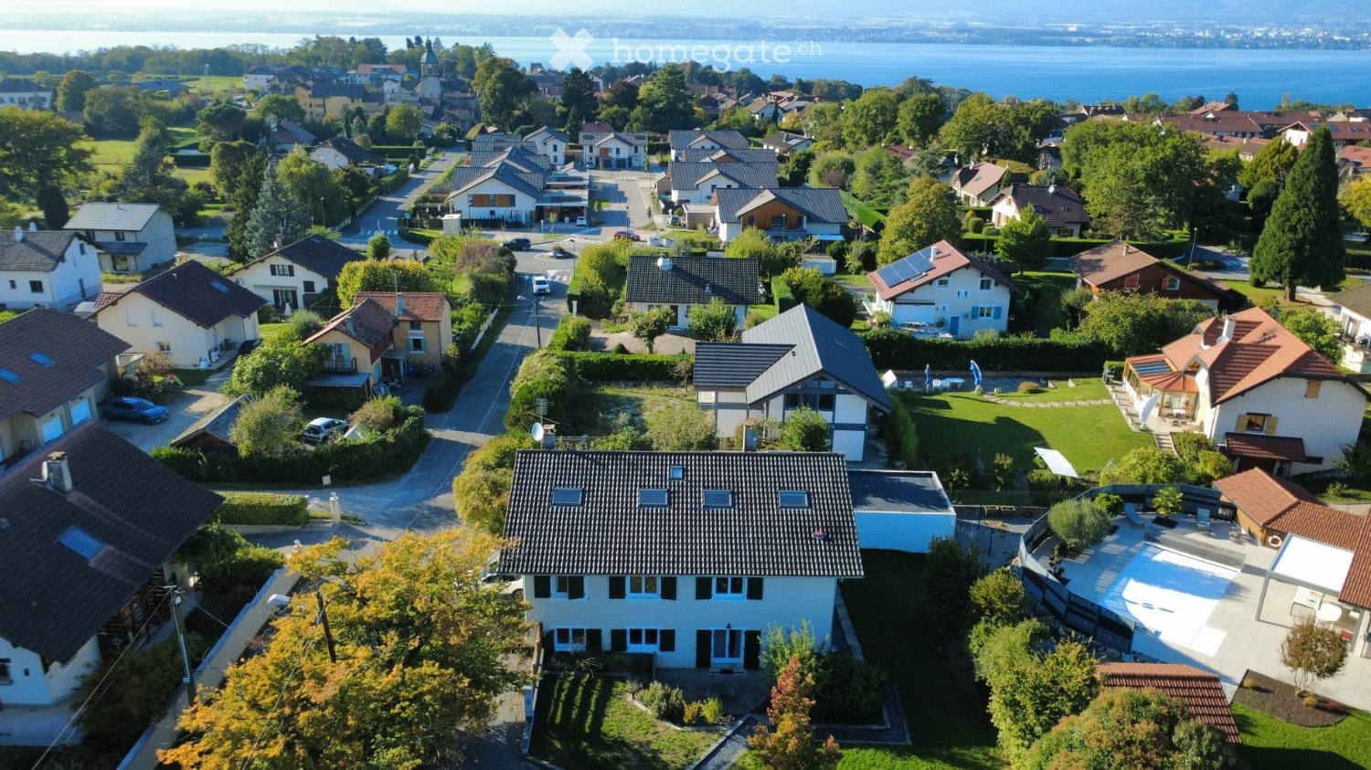 Residential neighborhood with multiple houses, varied architectural styles, some with solar panels, one with a swimming pool, green lawns, trees and bushes, distant view of water and hills.