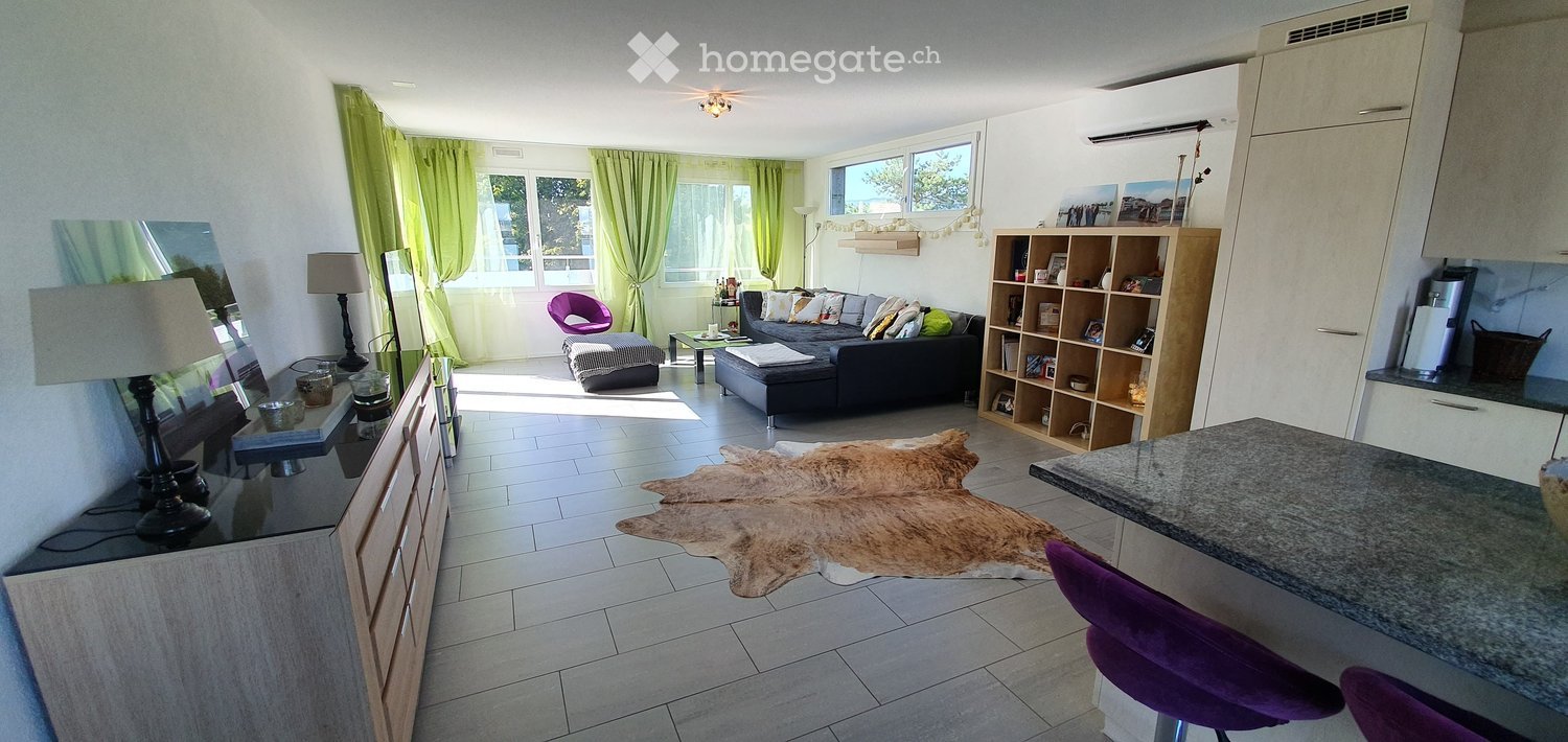 Open floor plan, black couch, wood flooring, two ottomans, wooden rug, wooden cabinet, grey counter top, purple chairs, bookshelves, green curtains