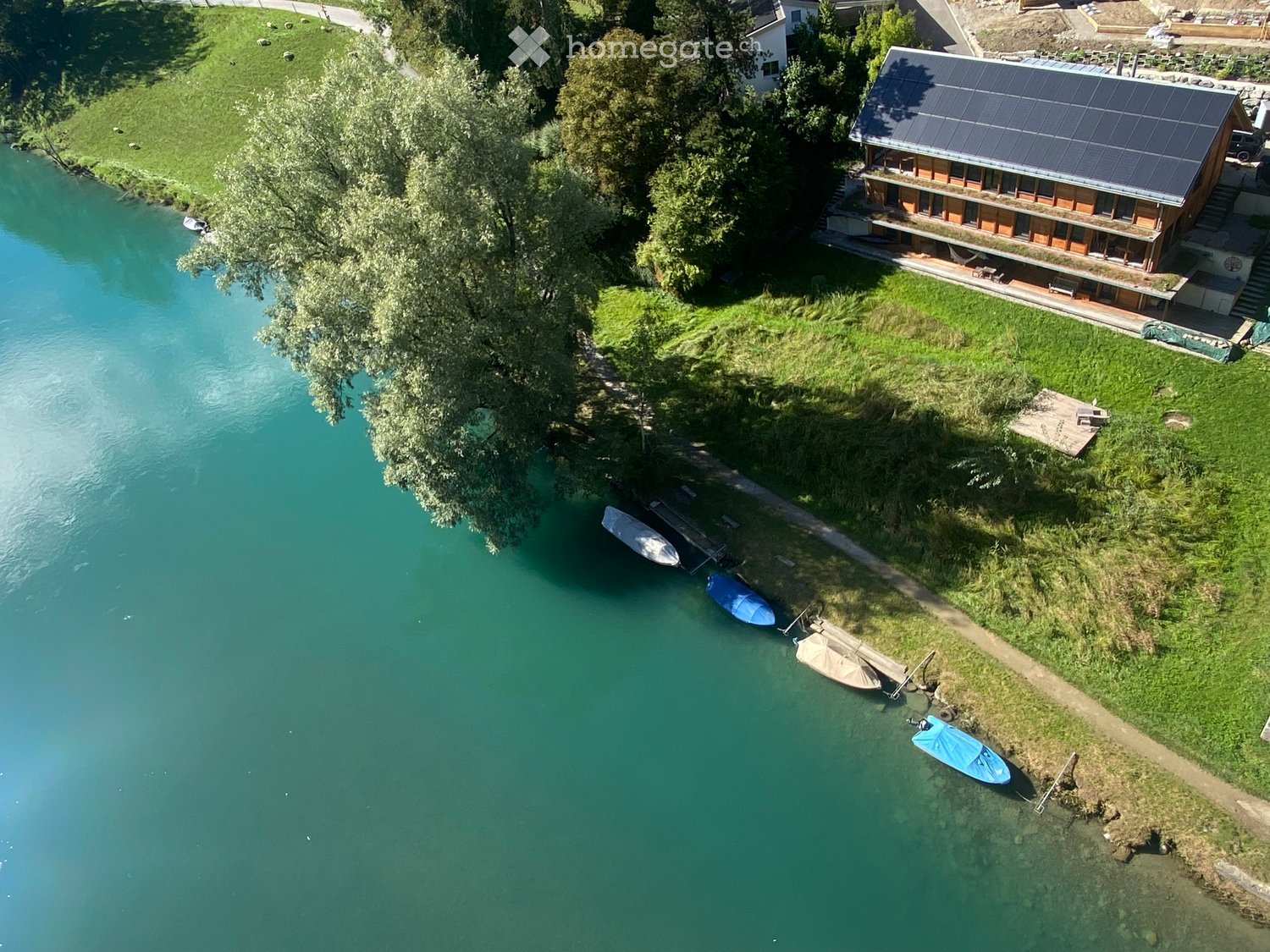 House by the water with a solar roof, close to a forest, boats docked.