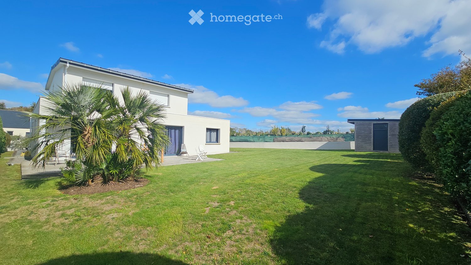 Modern house, white exterior, tiled roof, large well-kept garden, two palm trees, outdoor seating area, two white chairs, paved pathway, shrubs, open field in the background.