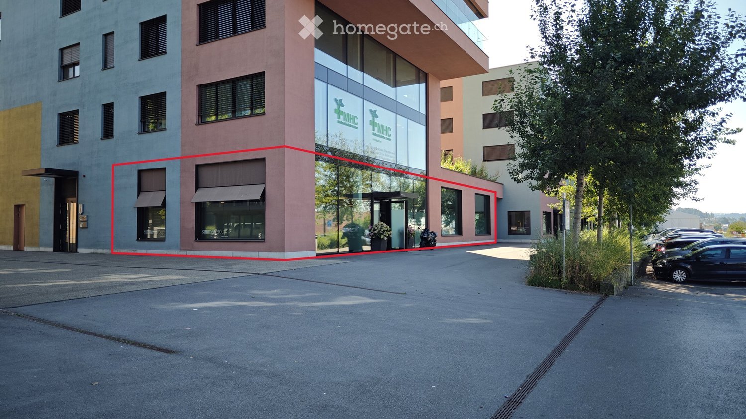2 story building, glass windows, red marking, entrance, several windows, 2 flower pots, cars parked, several trees