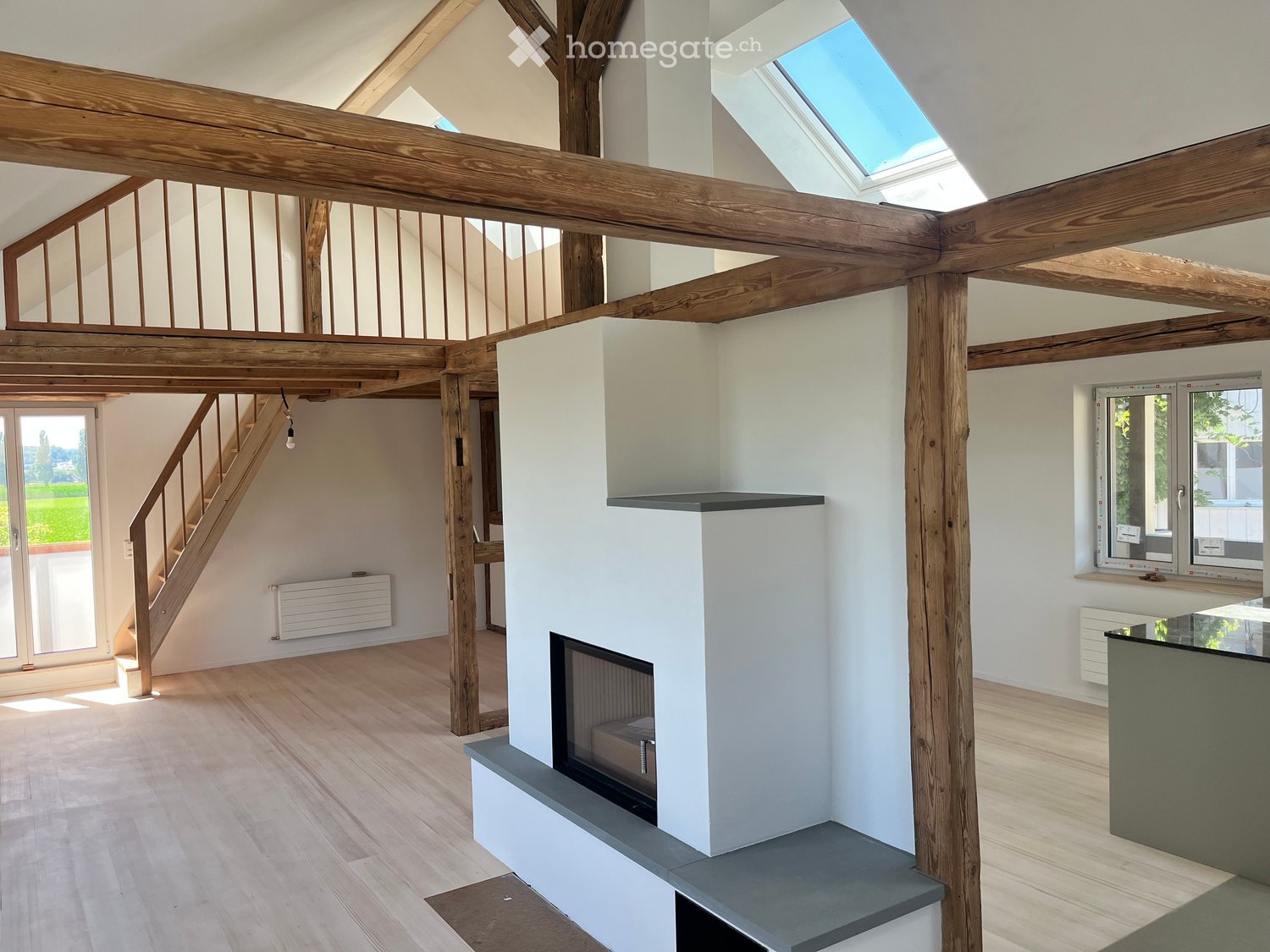 Open-concept living space with exposed wooden beams, a modern fireplace, and large windows providing natural light. The room has a minimalist and contemporary design with a neutral color palette.