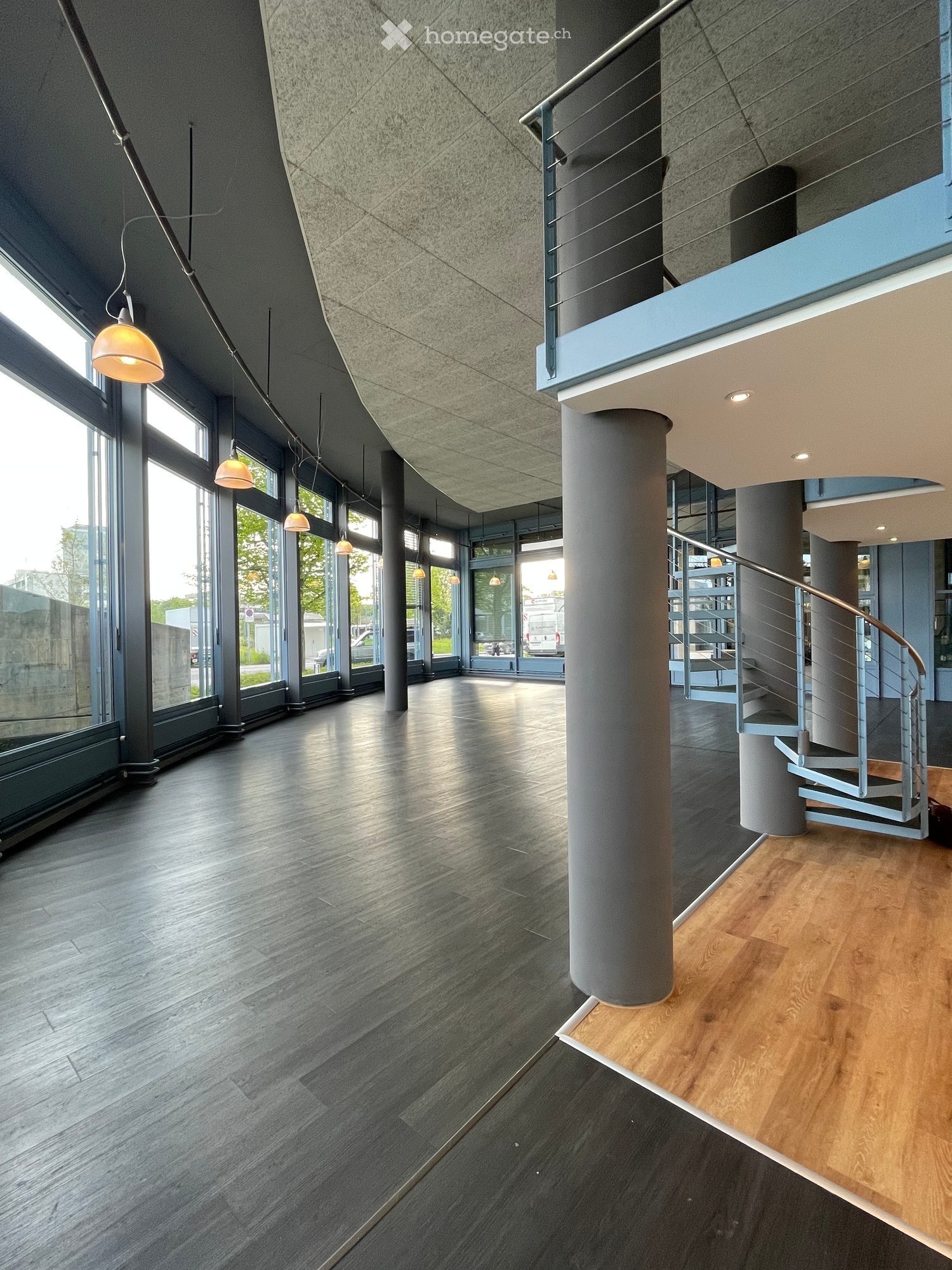 Spacious open room, with large windows and gray walls, metal railings, metal lamp-shades, wooden floors and spiral stairs.