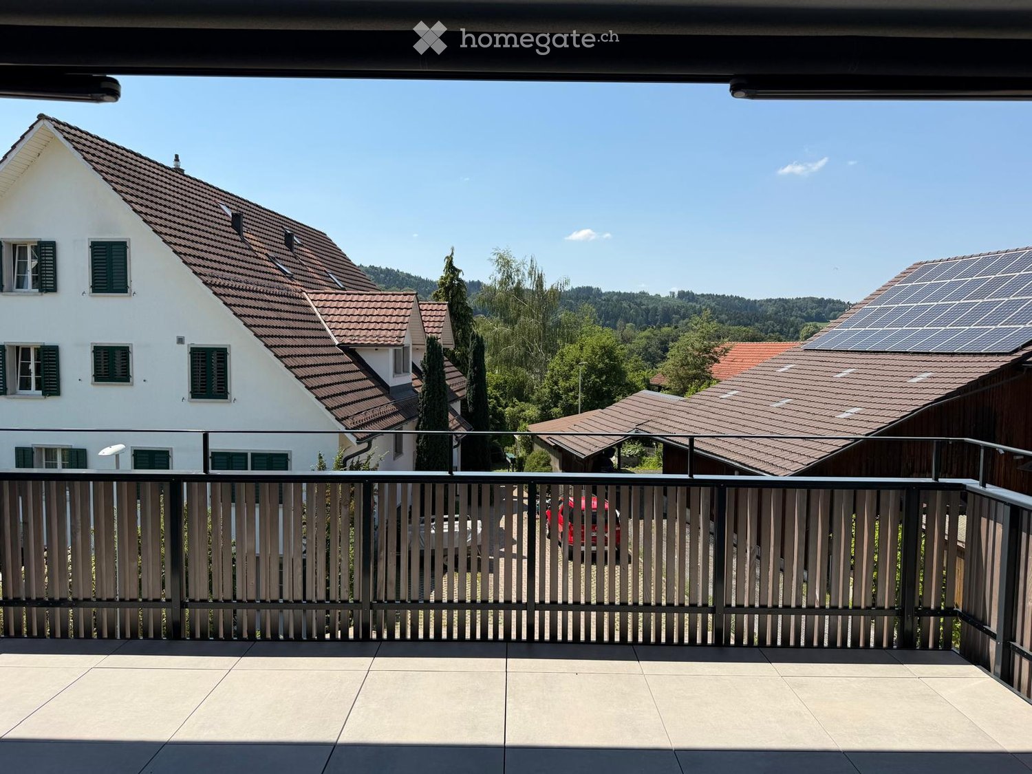 Balcony with tiled floor, metal railing, overlooking a residential area with houses, trees, and solar panels on one roof.