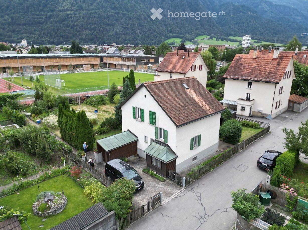2 story house with brown roof, green shutters, and a small attached building, garden with plants and cars parked in front