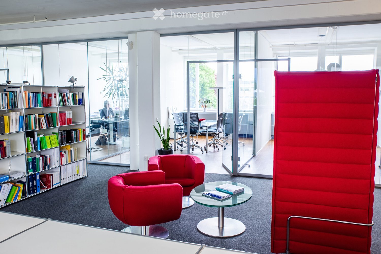 modern office space with red chairs and tables, large windows, shelves filled with books, a plant in the corner, an office cubicle