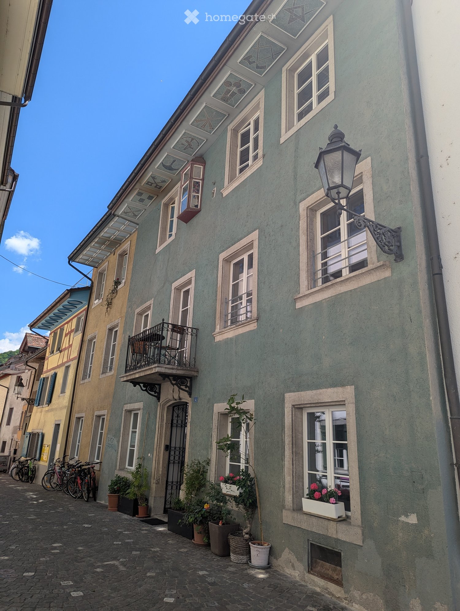 Two story building with balconies and windows, multiple plants outside, cobblestone road