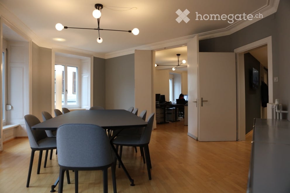 A large meeting room with a long table, a gray table, and several chairs. In the corner, a TV is mounted on the wall. Windows are present on the left side. Two people are working on the computer at the end of the room.