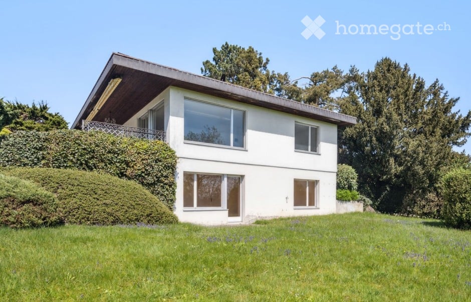 Single-story house, white exterior, brown roof, balcony, large windows, surrounded by a lush green lawn and hedges