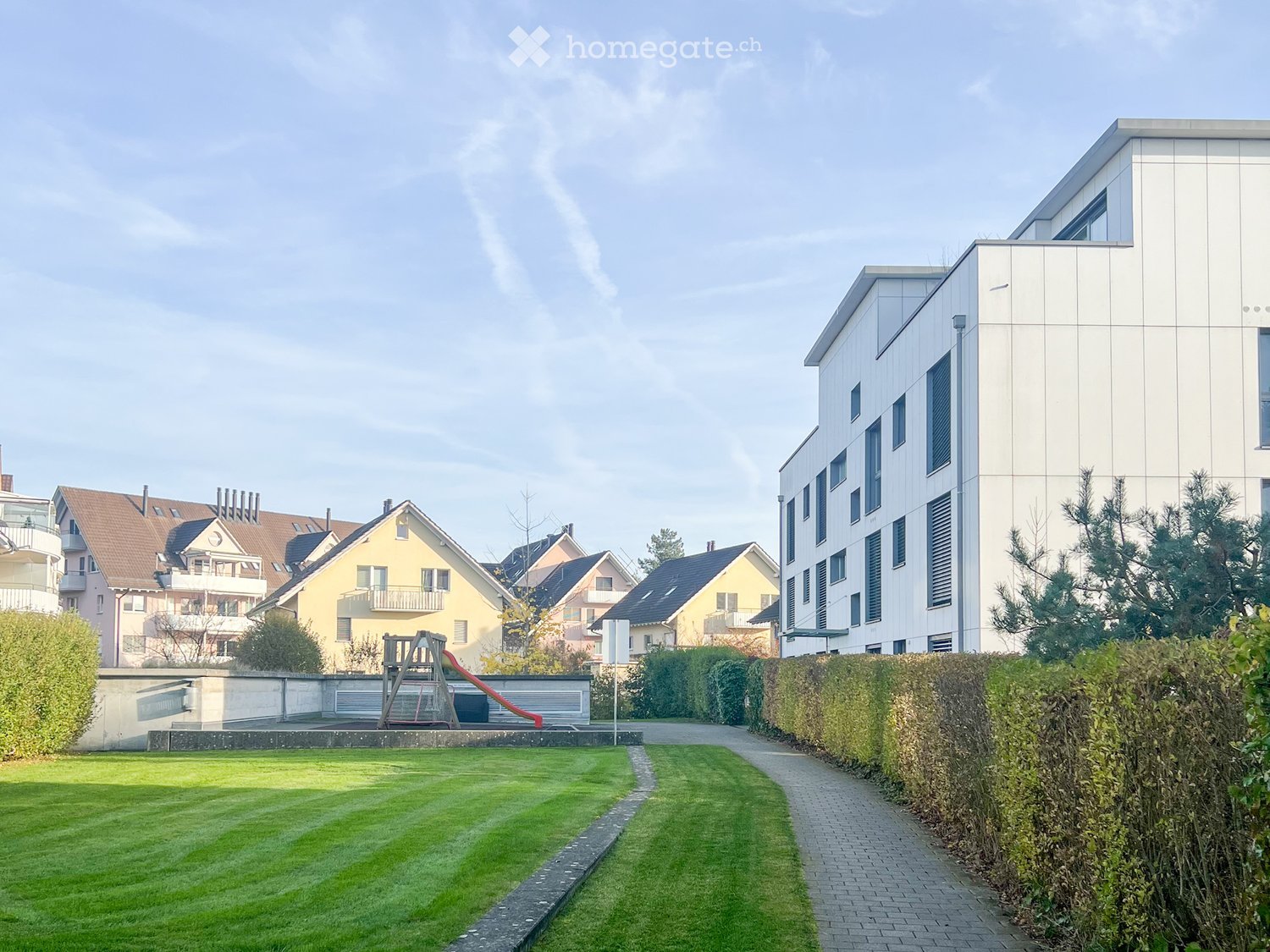 Multi-story residential building, green lawns, walkway, hedges, and playground with a slide.