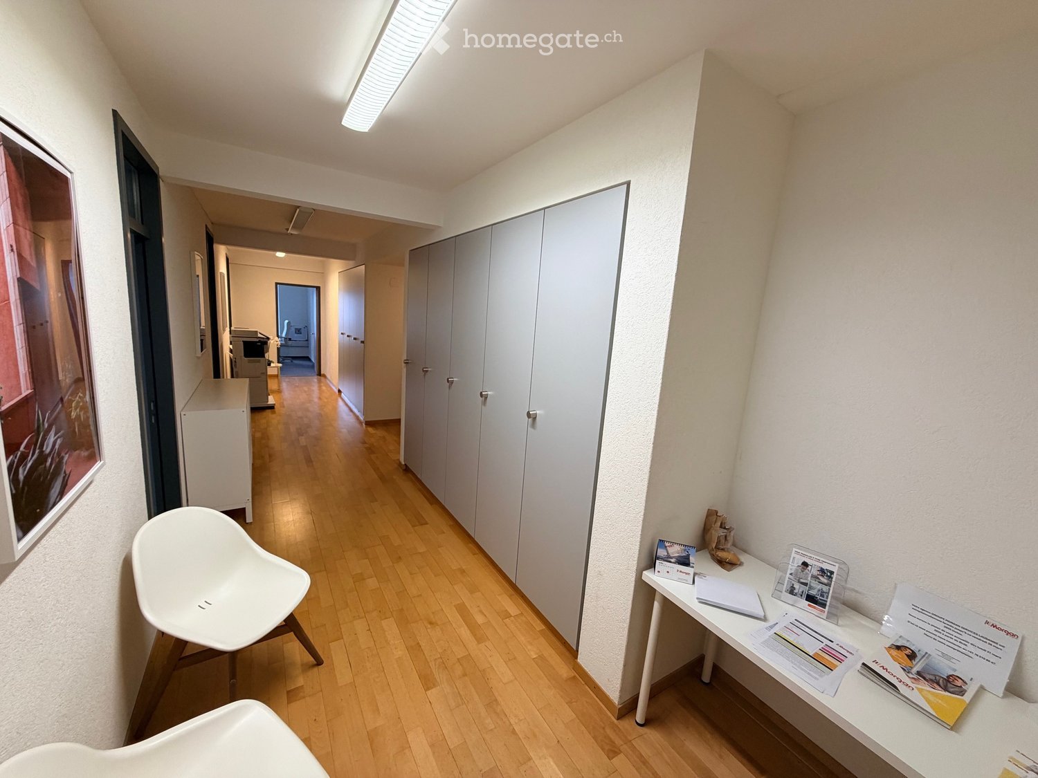 A hallway with a wooden floor, white walls, and cabinets, white chairs, a desk with brochures, a picture, and other items