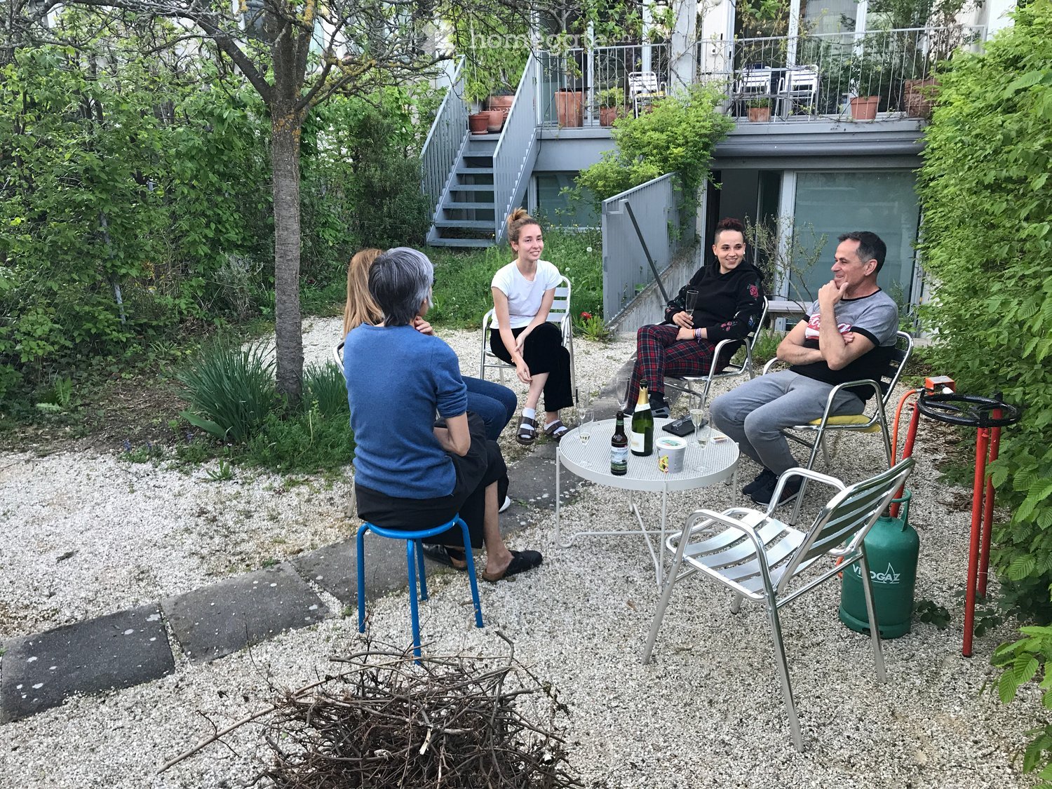 a group of people sitting around a table, in front of a building, trees and bushes