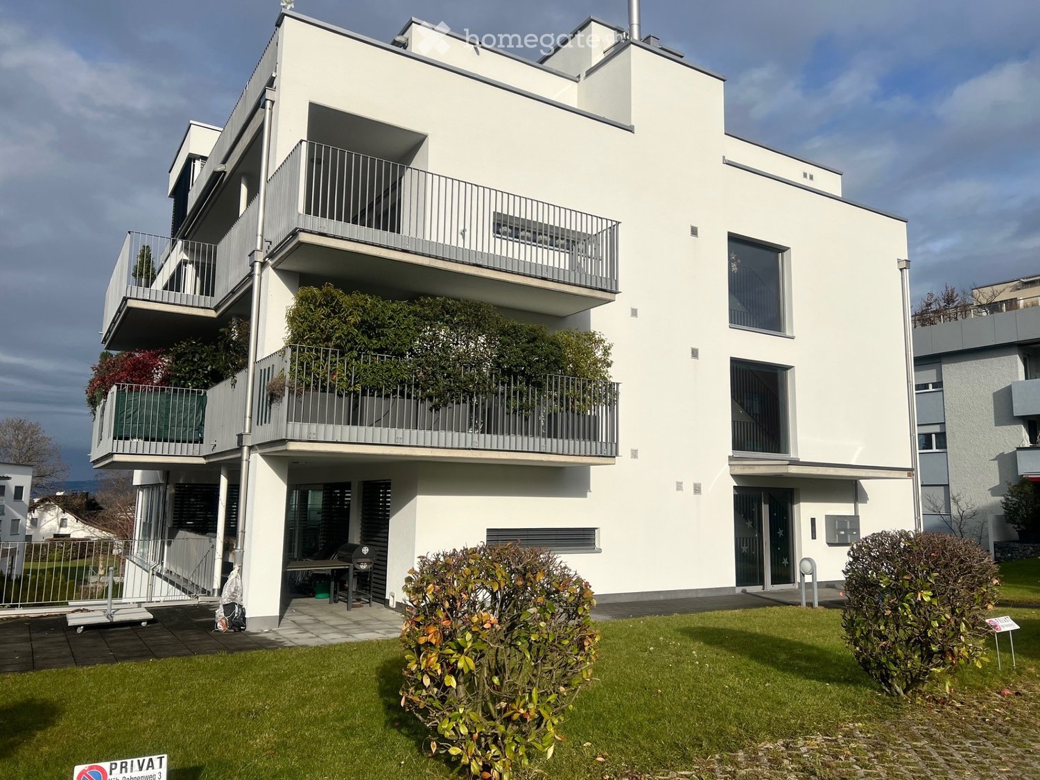 Modern white apartment building with multiple balconies, raised ground floor, and well-maintained green lawn in front.