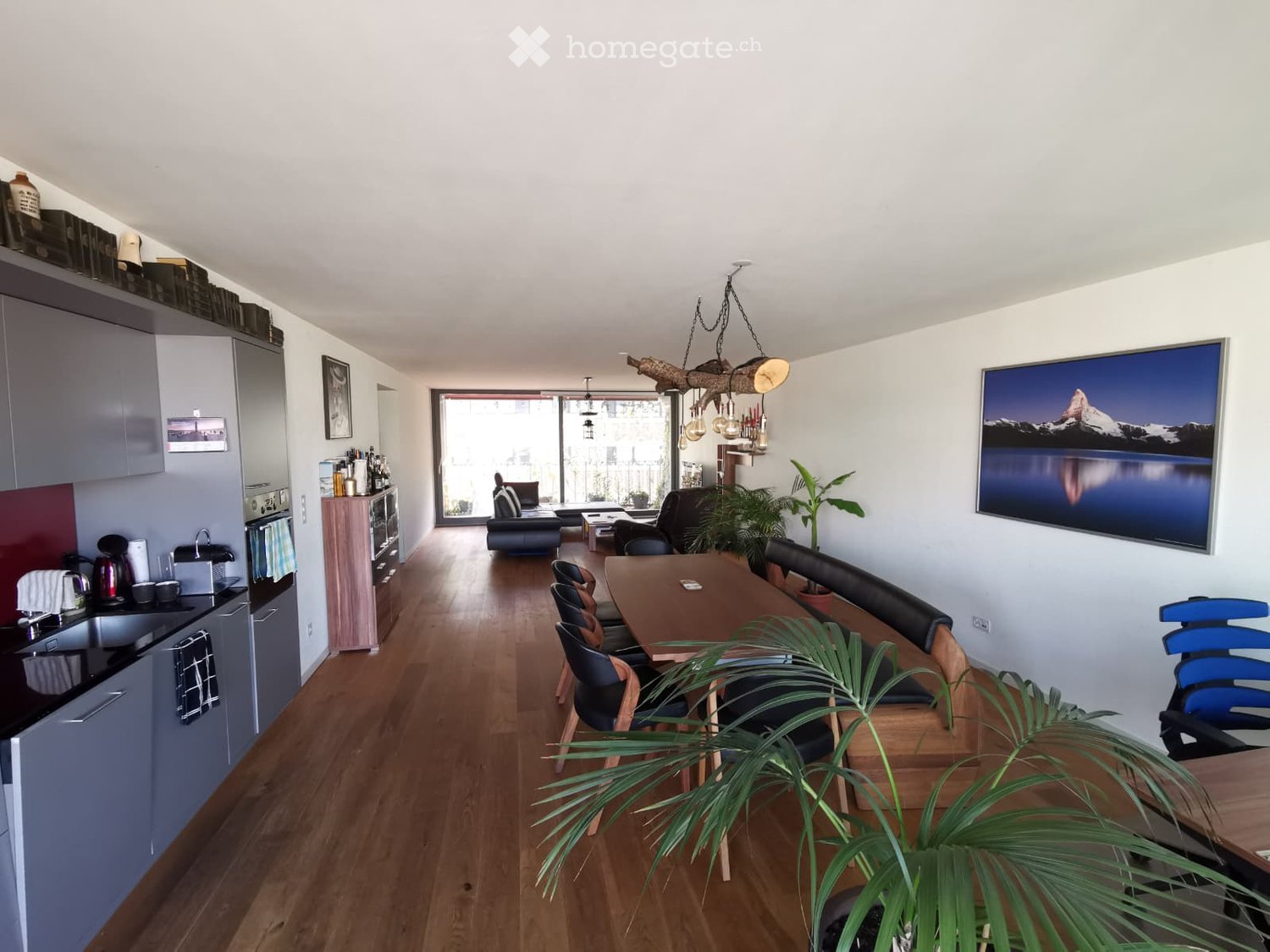 open floor plan, wooden floor, white walls, kitchen with cabinets, dining table with chairs, living area with couch, large window with balcony, framed picture