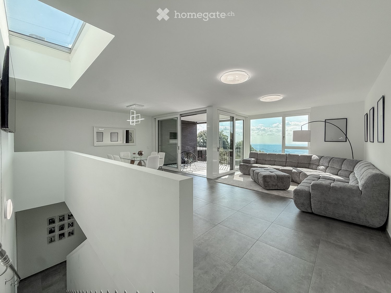 Modern living space, light grey floors, large windows, L-shaped grey couch, central island dining table, balcony with glass doors, large skylight, open kitchen with a glass wall, ocean view, tiled flooring, white walls, built-in shelves, ceiling-mounted l