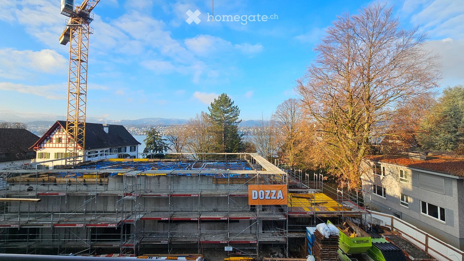 Building under construction, with crane, scaffolding, several houses visible in background, trees, mountains, lake in the distance.