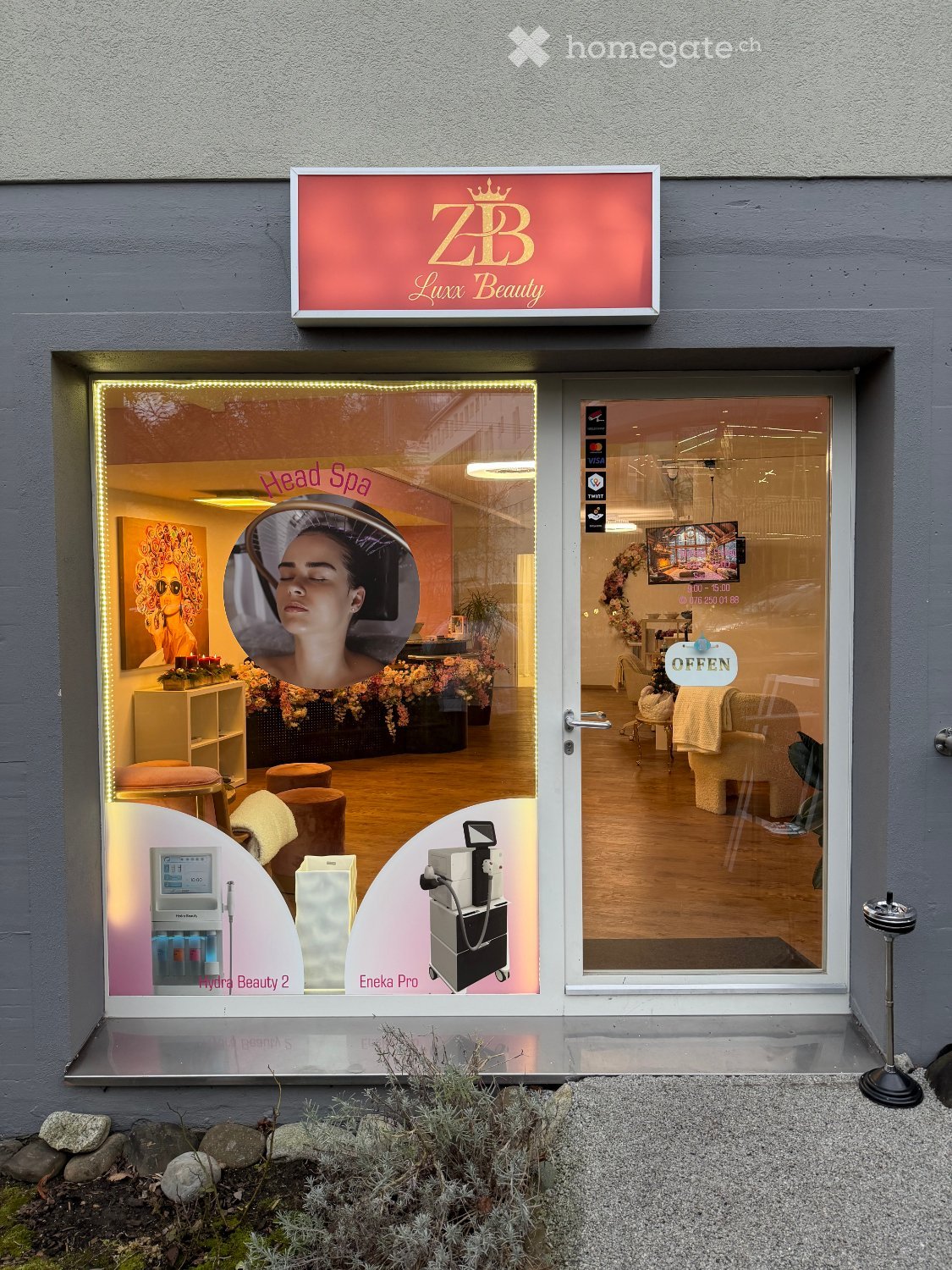 Facade of a beauty salon with large glass entrance doors and windows, illuminated red sign with 'ZB Luxx Beauty' text and logo, interior visible with spa equipment and seating.