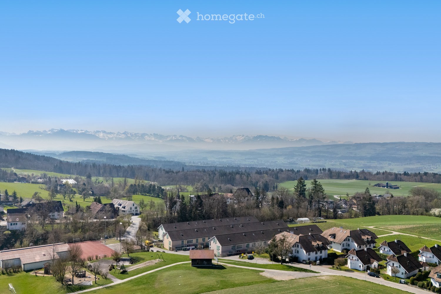 Group of detached houses with various sizes and architecture, surrounded by green fields and distant mountains