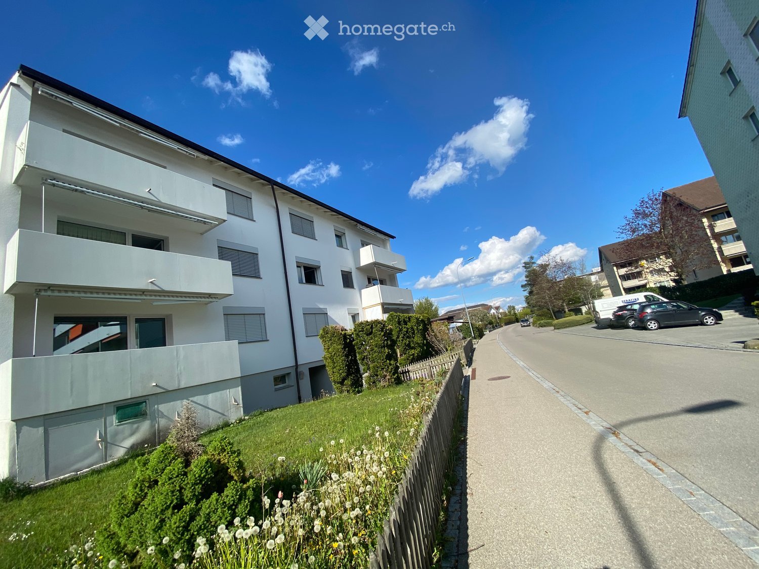 3 story building, white exterior, balconies, green lawn, parked cars, street