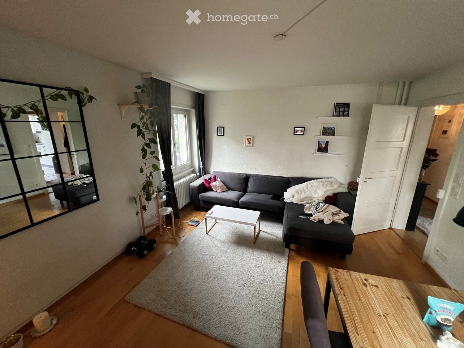 Large living room with gray sectional sofa, white coffee table, large rug, wooden dining table, radiator, shelves with framed pictures, mirror on wall.