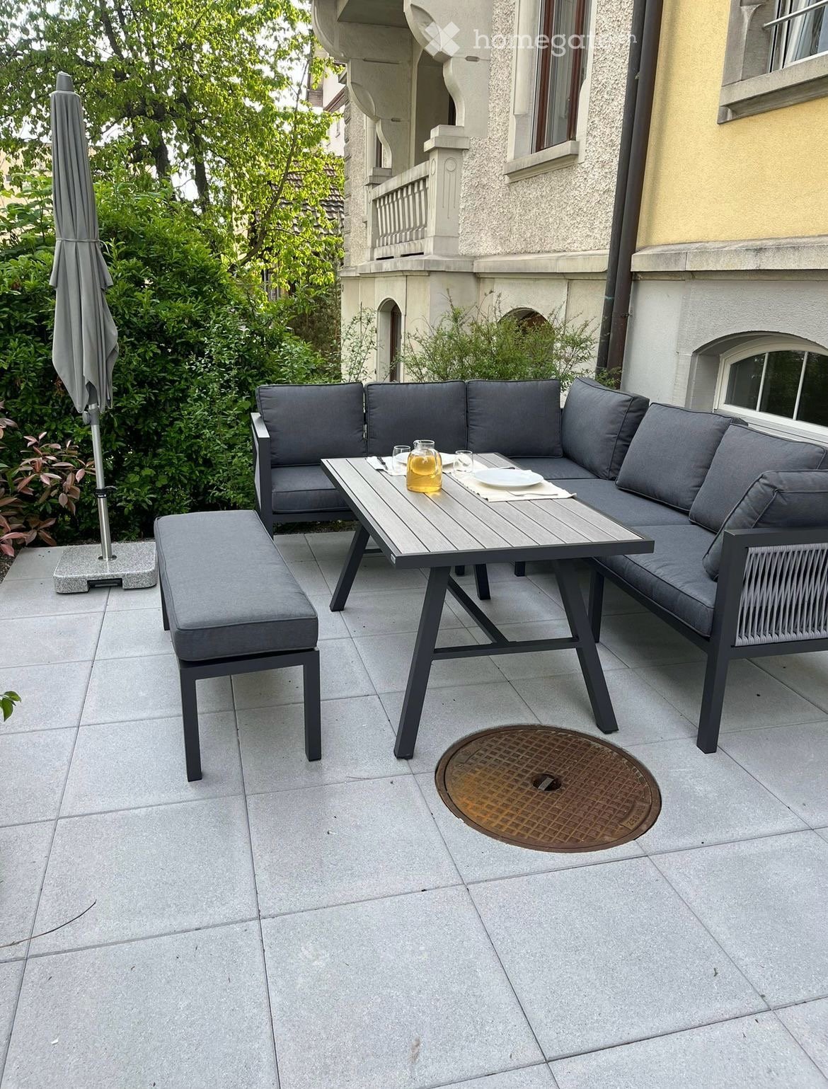 outdoor furniture set including sofa, bench, and table, metal table with glass top, grey cushions, grey umbrella, grey stone patio