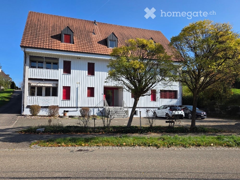White painted house, brown roof, red shutters, cars parked in front, trees, paved driveway
