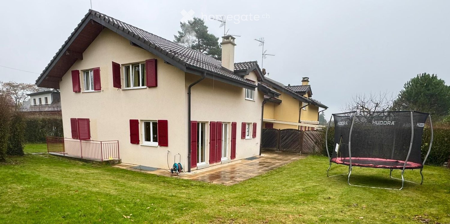 House with gable roof, painted white and beige, red shutters, tiled path, fence, trampoline, large garden