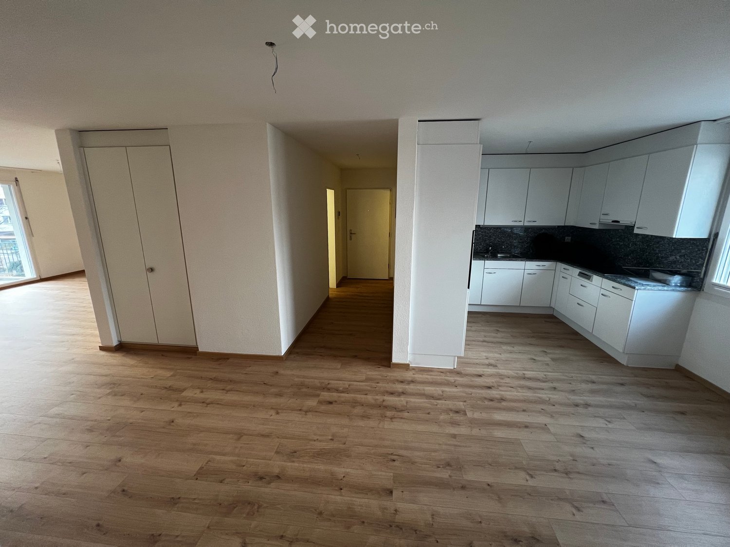 Empty room with wooden floors, white cabinets, white walls, glass window, hallway, kitchen