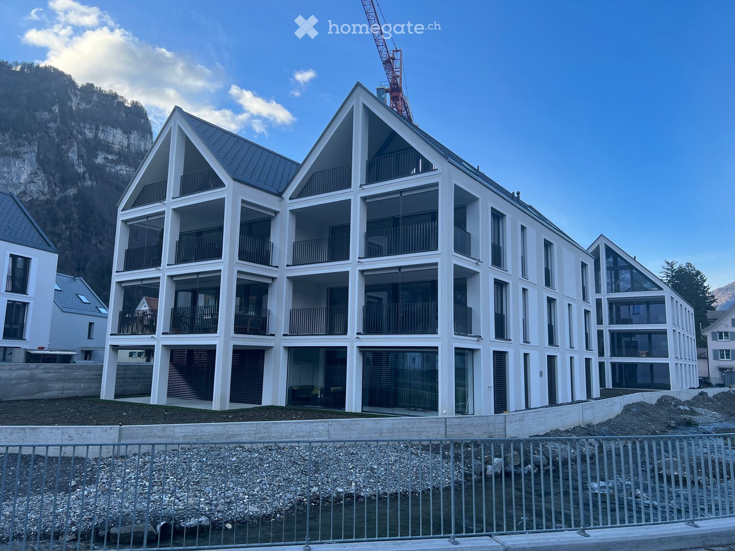Large multi-family residential building, white exterior, large windows, multiple balconies, construction area nearby, mountains in the background.