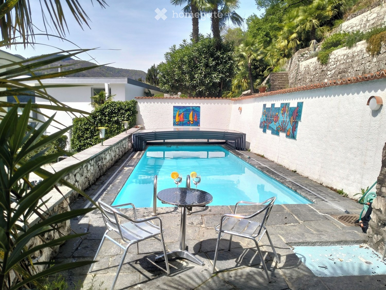 An outdoor area with a swimming pool, two metal chairs, a round table, two glasses with lemon slices, and art decorations on the white wall.