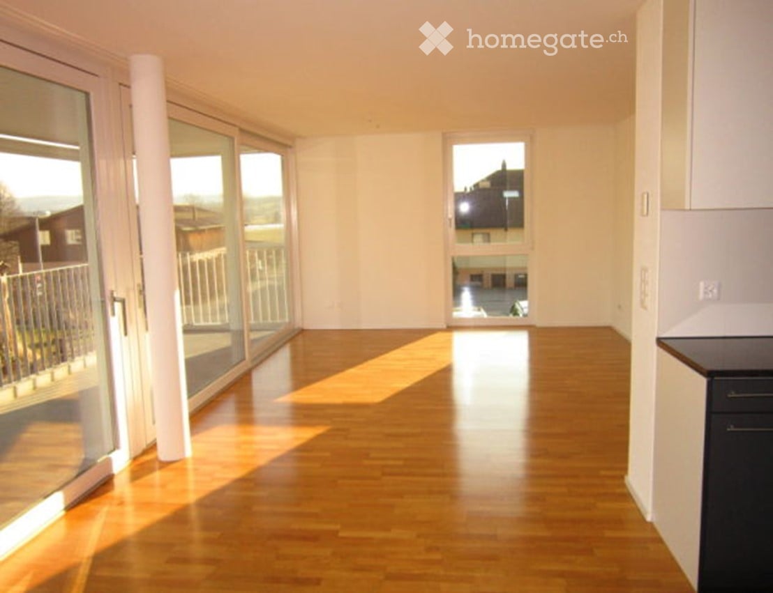 An empty room with wooden floors, white walls, large glass sliding doors, balcony, and a view of a street with a building.