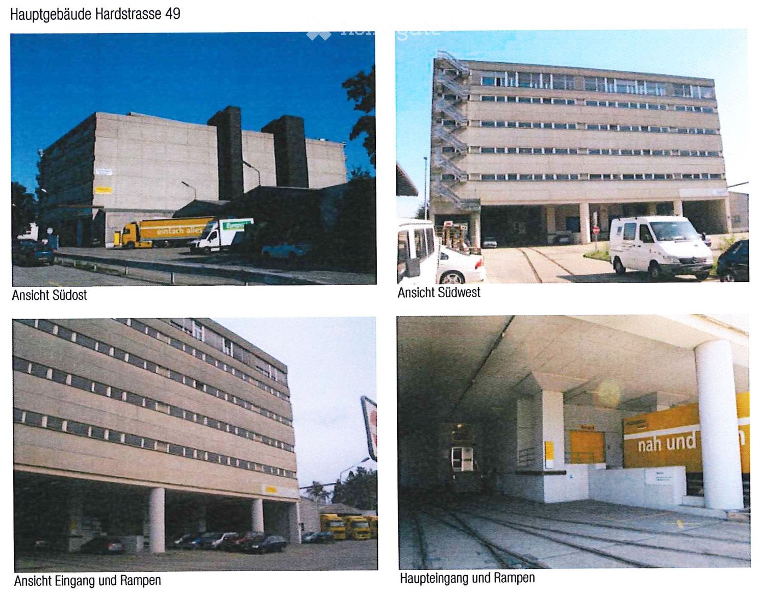 Large multi-story building with numerous windows, multiple trucks parked outside, open area with ramps and train tracks inside, several cars parked in a lot in front of the building.