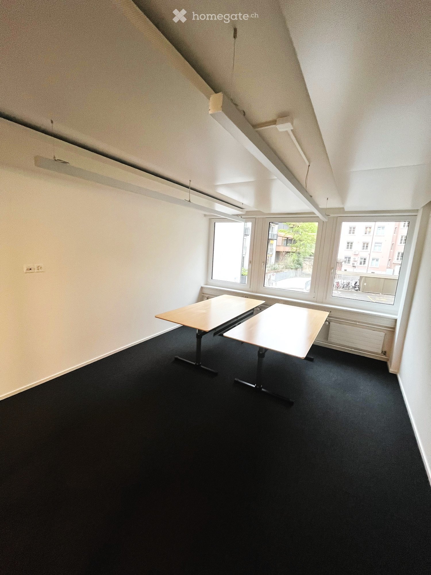 Empty room with two tables, white walls, black carpet, and windows facing the street