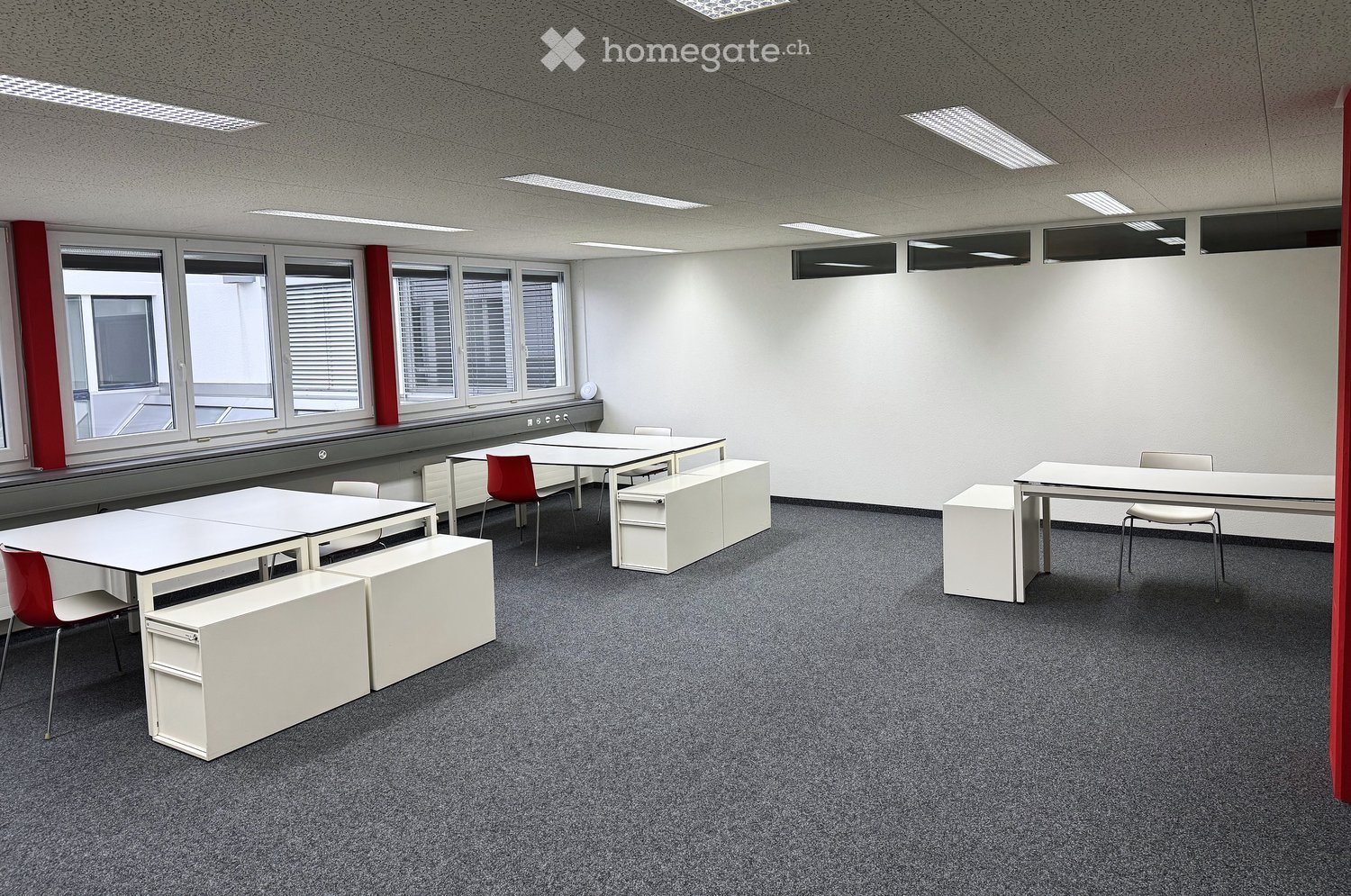 Empty room, white walls, carpet flooring, desks and chairs, windows with blinds, ceiling lights
