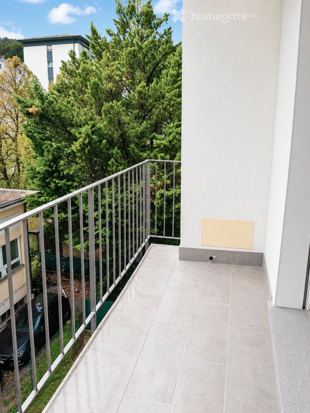 Balcony with grey tiled floor, metal railing, and a view of green trees and neighboring buildings.