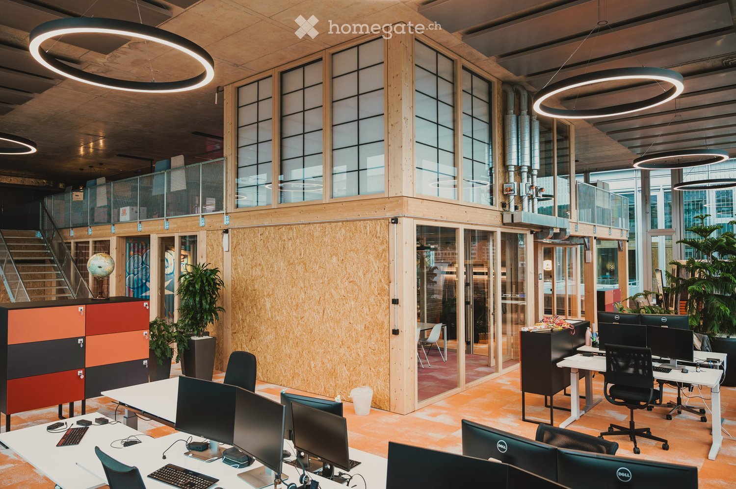 A modern open-plan office with a contemporary design, featuring an array of desks, chairs, and computers. The walls are partially made of wood and glass, providing a bright and spacious feel. Circular lamps hang from the ceiling, and the floor is covered