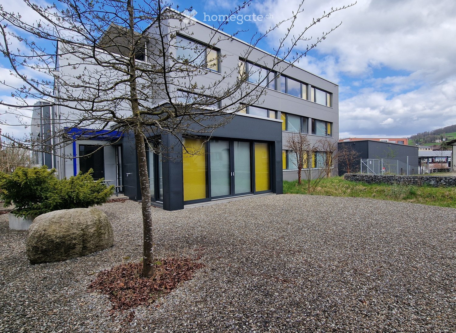 Modern two-story building with grey and yellow facade, large windows, and a gravel driveway.