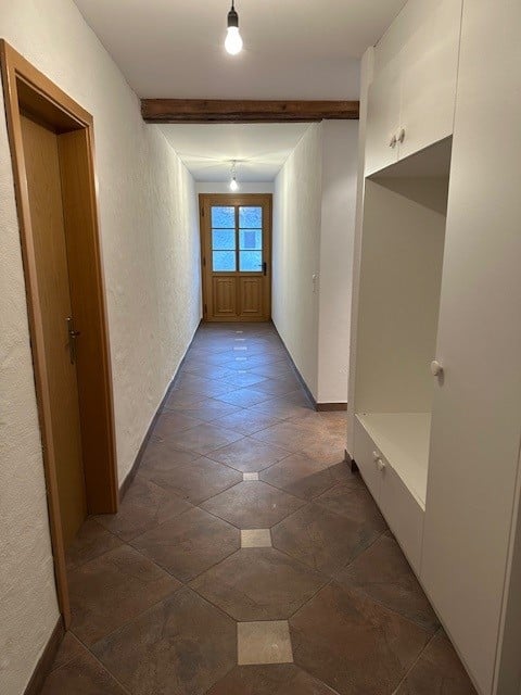 Empty hallway, tiled floor, wooden doors, white walls, ceiling lights, window with glass