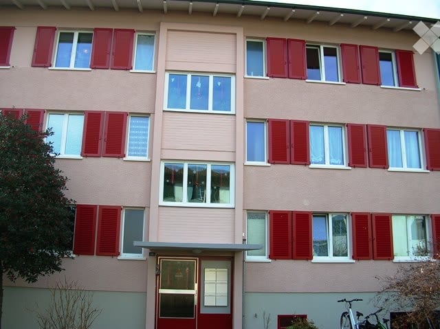 A five-story building with red shutters, glass windows, and a red door on the ground floor. There are bicycles parked in front of the building and two plants in front.