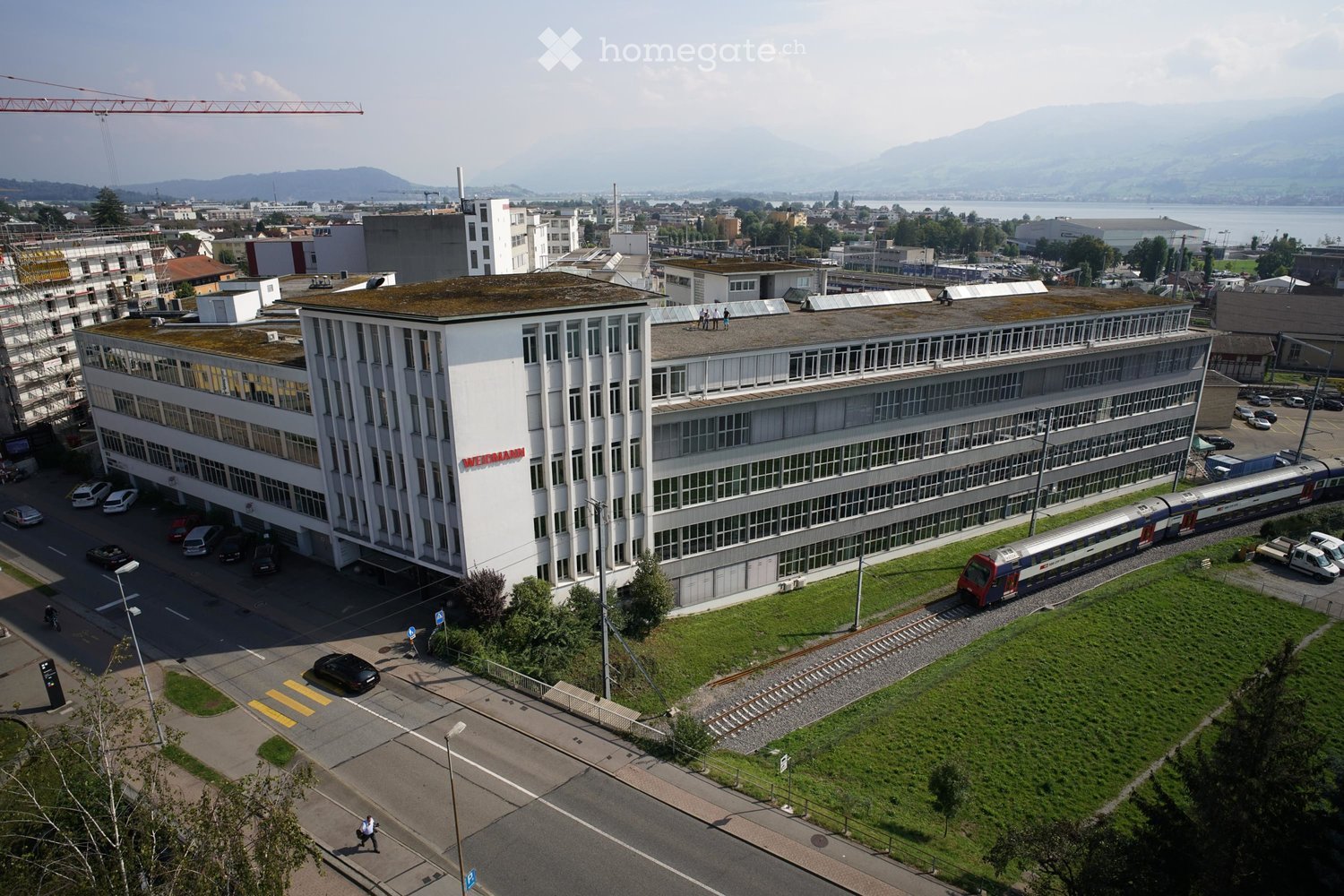 Industrial building, modern architecture, large, white structure with many windows, red signage, outdoor parking, railway track in front, mountain view, urban area.