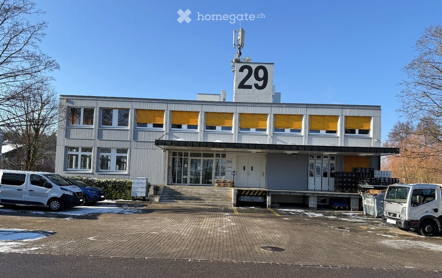 A gray building with a flat roof and yellow shutters on the windows, with a number 29 at the top, featuring a large parking area with vehicles parked in front.