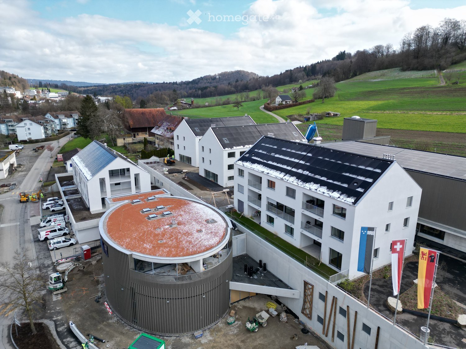 Aerial view of a modern building complex with a circular structure, surrounded by a green field and mountains in the background.