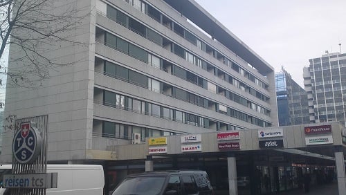 A gray building with many windows and a lot of signs. Below the building is a parking area. There are two cars parked in front of the building.
