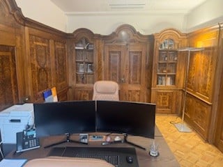 an office desk with two monitors, a phone, keyboard and mouse on top, a wooden cabinet with books, a chair, a lamp
