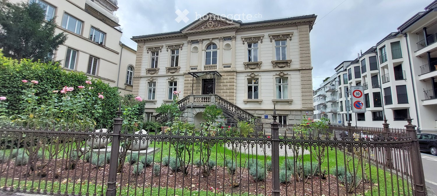 Three story brick house, many windows, grand entrance, ornate details, garden with flowers and greenery.