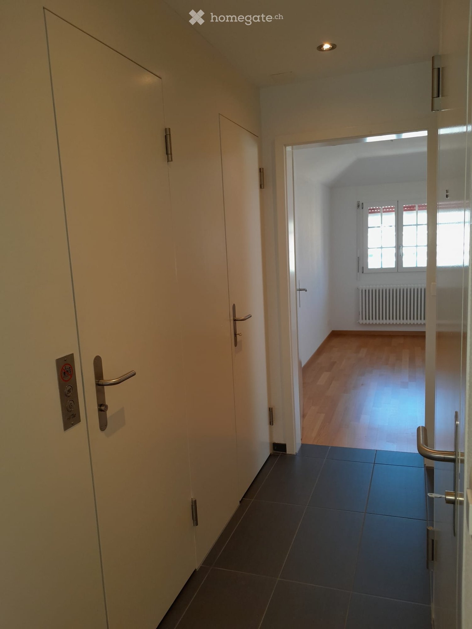 Empty corridor with wooden floors, white walls, glass doors, and a window with a radiator.