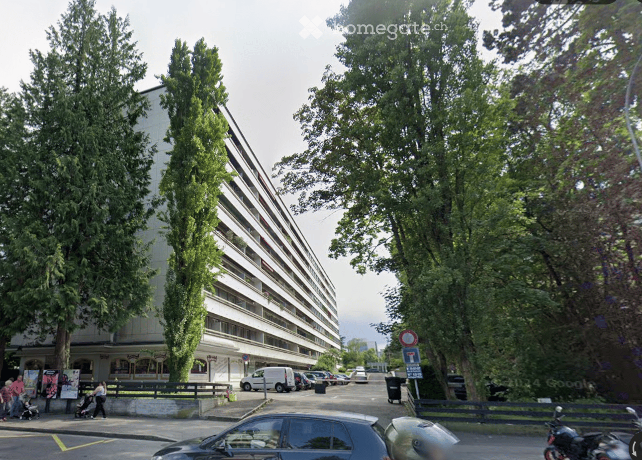 Apartment building, modern architectural design, multiple balconies, surrounded by trees and parking area, street facing facade, ground floor shops