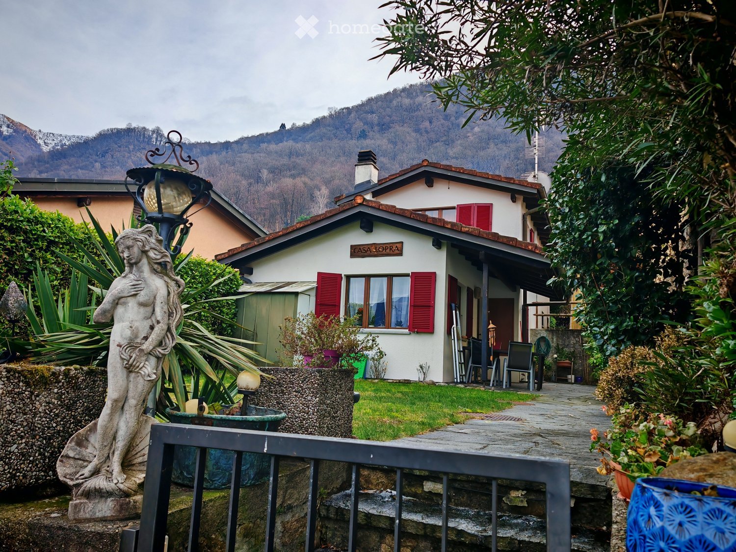 White house with red shutters, two story with a red tiled roof, sitting in a garden with decorative plants and a statue, mountains in the background