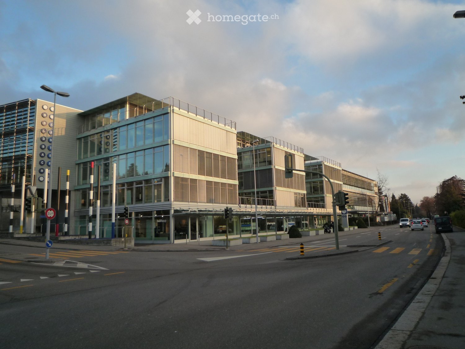 Modern multi-story building with large glass windows and a flat roof, located at a street corner with traffic signals and road markings.