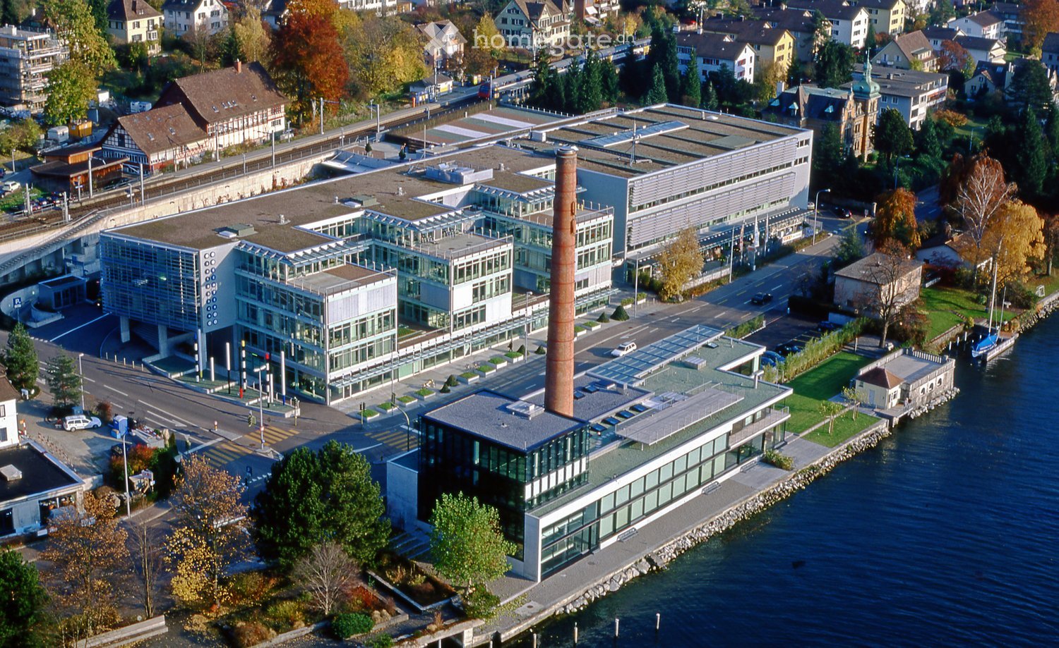 Aerial view of a modern industrial complex with large glass windows, a prominent chimney, and surrounding urban area with residential buildings, trees, and a waterfront.